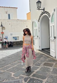 Conjunto de duas peças em castanho claro com um top crop sem mangas e uma saia longa; camisa xadrez vermelha atada na cintura; botas pretas até ao tornozelo.