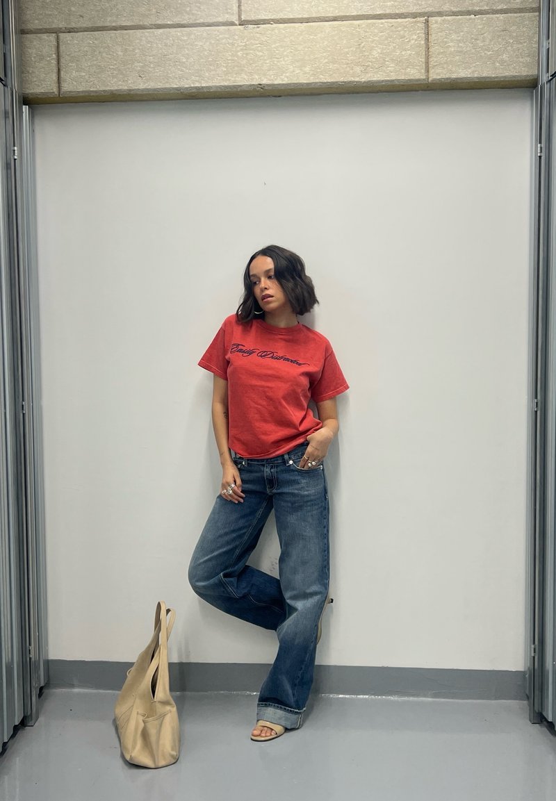 Donna con capelli corti e scuri che indossa una t-shirt rossa con grafiche, jeans blu e sandali, appoggiata a un muro bianco con una borsa beige sul pavimento nelle vicinanze.