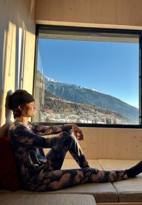 Μακρυμάνικη, εφαρμοστή ρούχα λούνας σε σκούρο tie-dye σχέδιο. Ο ιστός φαίνεται να είναι μαλακός. Καθισμένος σε ένα φωτεινό δωμάτιο με θέα βουνό.