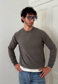 Grå strikket trøje med rund halsudskæring, parret med en hvid lagdelt kant og blå denimjeans, iført sølvringe og store blåtonebriller.