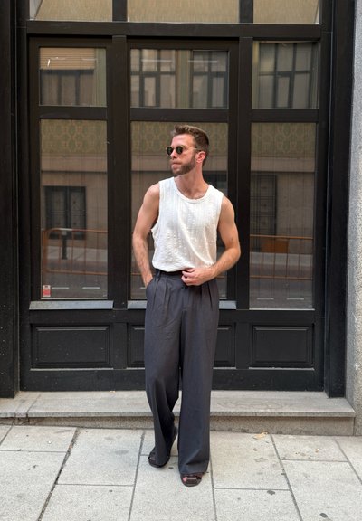 Top sin mangas blanco con detalles texturizados, pantalones de pierna ancha gris oscuro y gafas de sol redondas, de pie frente a una ventana con marco negro.