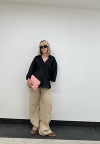 Chemise noire surdimensionnée à boutons, pantalon beige à jambes larges, pochette matelassée rose et lunettes de soleil foncées devant un mur blanc. Chaussures marron avec lacets blancs.