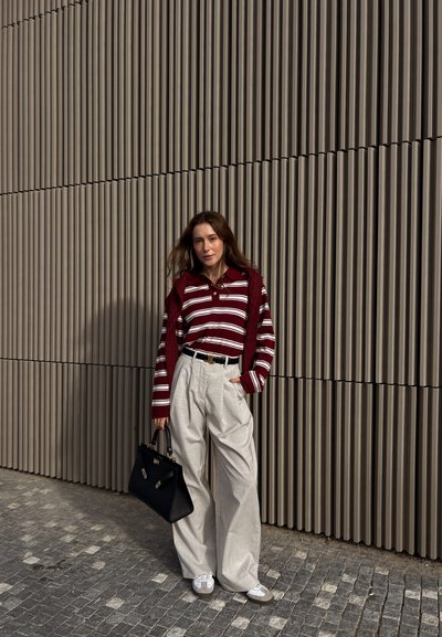 Camisa de rayas marrón y blanca de manga larga, combinada con un suéter marrón, acompañada de pantalones beige anchos y un bolso negro. Fondo texturizado.