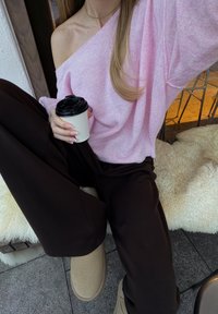 Personne portant un pull rose décolleté et un pantalon foncé, assise sur un banc rembourré, tenant un gobelet de café à emporter.