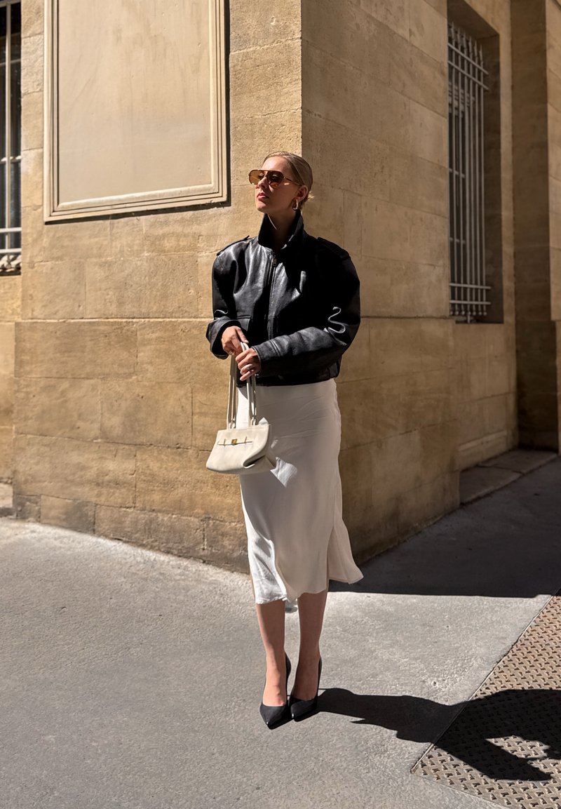 Black leather bomber jacket, white satin slip dress, black stiletto heels, and small off-white handbag; sunglasses complete the look.