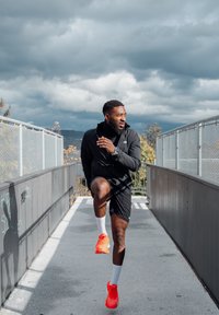 Man in black sportswear and bright orange shoes warming up on a fenced outdoor path under a cloudy sky.