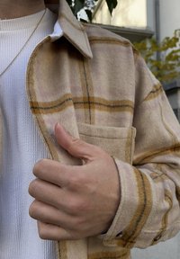 Camicia di flanella beige con motivo a quadri giallo e rosa chiaro, dotata di taschino al petto e polsini con bottoni, indossata sopra un tessuto bianco a coste.