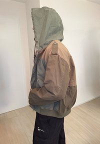 Personne portant une veste à capuche marron et grise, capuche relevée, mains dans les poches, debout à l'intérieur contre un mur blanc.
