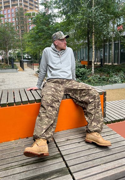 Sudadera gris, pantalones de camuflaje y botas beige. Estilo casual con un ajuste relajado. Banco de madera y naranja en un entorno verde al aire libre.
