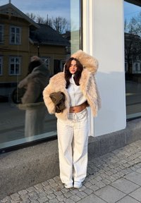 Beżowa kurtka z sztucznego futra, jasnoszara bluza z kapturem na zamek, białe szerokie spodnie, trzyma brązowy sweter z fakturą, stoi obok odbijającej powierzchni szklanej.