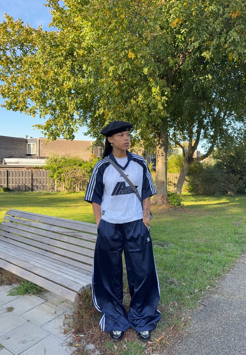Zwarte baret, wit en marineblauw sportshirt met logo, en marineblauwe wijde trainingsbroek. Glad materiaal en drie strepen op de mouwen en broek.