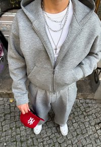 Szürke zipzáras kapucnis pulóver és passzoló melegítőnadrág, fehér bordázott póló, rétegezett ezüst nyakláncok, piros NY sapka és fehér sportcipők kiegészítőkkel.