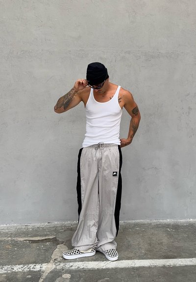 Pantalones de chándal grises y negros con un diseño ligero, usados con una camiseta sin mangas blanca y zapatillas de cuadros. Los accesorios incluyen gafas de sol y un gorro negro.