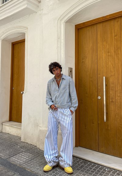 Camisa de rayas en azul y blanco, pantalones de rayas holgados y calzado amarillo. De pie contra una pared blanca con puertas de madera.