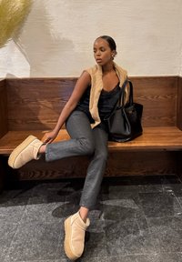 Femme assise sur un banc en bois, portant un top noir, un gilet beige, un pantalon gris et des bottes beiges, ajustant une botte avec un sac à main noir à ses côtés.