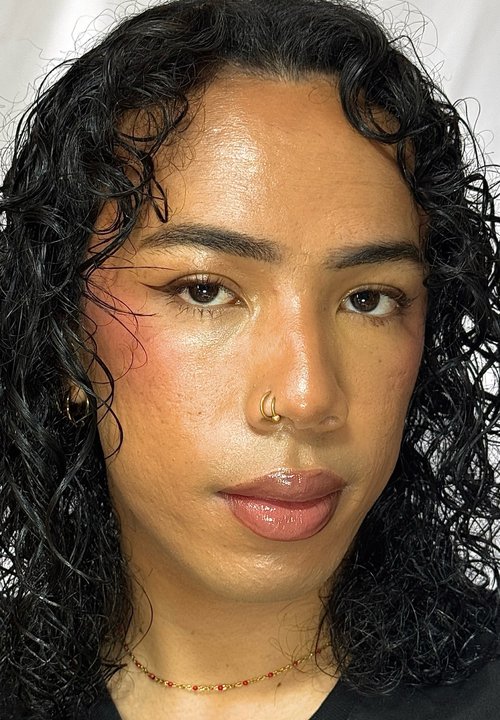 Person mit lockigem Haar, die blaue und silberne Lidschatten mit kleinen Strasssteinen trägt, neutralen Lippenstift und einen goldenen Nasenring.