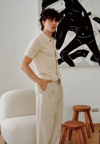 Camisa de manga curta bege clara com riscas verticais, combinada com calças plissadas bege claro. Textura suave, sentado numa cadeira curva creme.