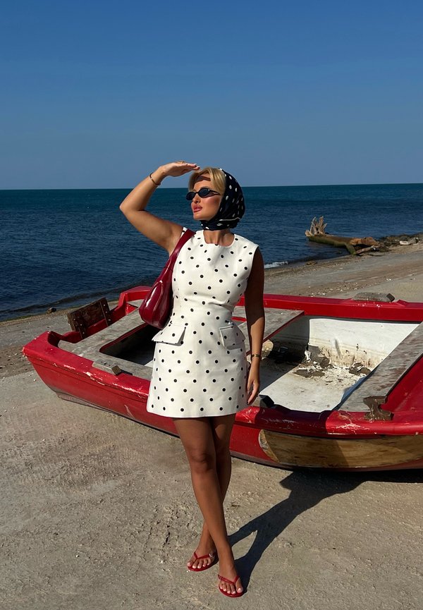 Femme en robe blanche à pois et foulard, protégeant ses yeux, debout près d'un bateau rouge sur une rive sablonneuse avec la mer et un ciel dégagé en arrière-plan.