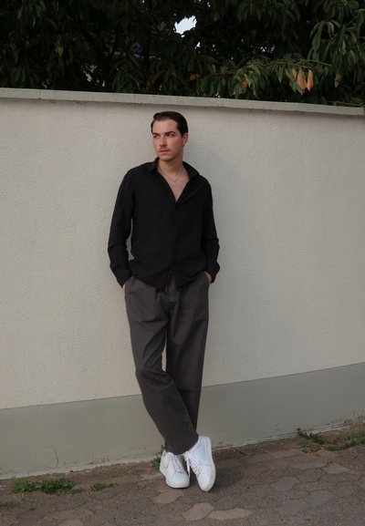 Camisa negra de botones, pantalones grises holgados y zapatillas blancas. De pie contra una pared clara con follaje verde arriba. Diseño casual.