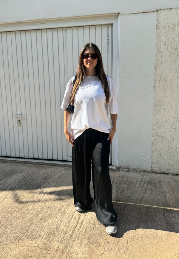 Joven mujer con el pelo largo y gafas de sol de pie afuera, lleva una camiseta blanca de gran tamaño, pantalones negros de pierna ancha y zapatillas.