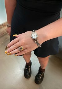 Srebrni i zlatni metalni sat s octagonalnim bijelim licem i detaljima od rhinestones, uparen s detaljnim zlatnim prstenom na ruci s dugim, dekorativnim noktima.