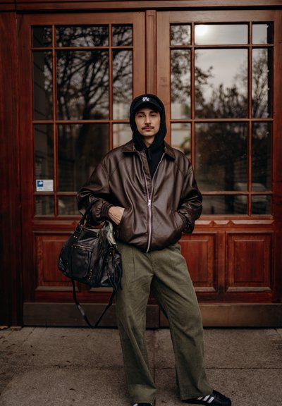 Joven con bigote que lleva una chaqueta de cuero marrón, sudadera con capucha negra, gorra negra, pantalones verdes y bolso negro, parado frente a una puerta de madera con paneles de cristal.