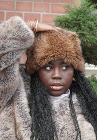 Fuskpälsjacka i grå nyanser med tät textur. Brun pälsmössa med mjuk finish. Accessoarer inkluderar smala guldsmycken.