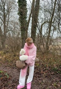 Pull rose clair en maille torsadée et écharpe, pantalon blanc, chaussures roses à lacets, tenant un sac à main beige. Entouré d'arbres nus et de feuilles.