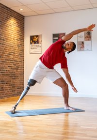 Man met een metalen prothese been in een yoga houding, gekleed in een rood shirt en witte shorts, op een blauwe mat in een studio met bakstenen muren.
