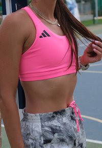 Žiarivo ružový športový crop top s čiernymi logovými detailmi, ktorý je kombinovaný so vzorovanými svetlošedými šortkami s šnúrkovým detailom.