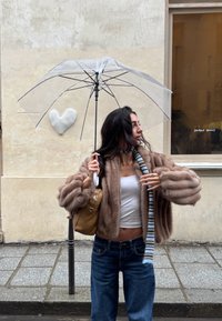 Femme en veste en fourrure tenant un parapluie transparent dans une rue de la ville, avec une œuvre murale en forme de cœur derrière elle.