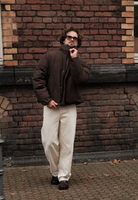Brown padded jacket with a high collar, beige wide-legged trousers, and dark shoes. Curly hair and glasses, standing against a brick wall.