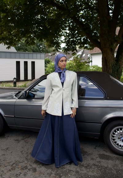 Chaqueta gris claro con bordado blanco, camiseta negra y falda maxi de mezclilla azul. El conjunto se complementa con un hijab azul y un anillo.