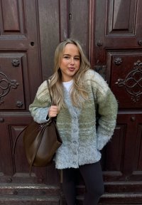 Ανοιχτό πράσινο και γκρι φουσκωτό πουλόβερ με κουμπιά, συνδυασμένο με μια καφέ δερμάτινη τσάντα. Σε φόντο μια σκοτεινή ξύλινη πόρτα.