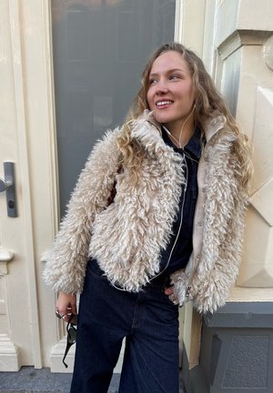 Mujer joven con una chaqueta beige esponjosa y pantalones oscuros, sosteniendo unas gafas de sol, de pie cerca de una puerta con auriculares puestos.