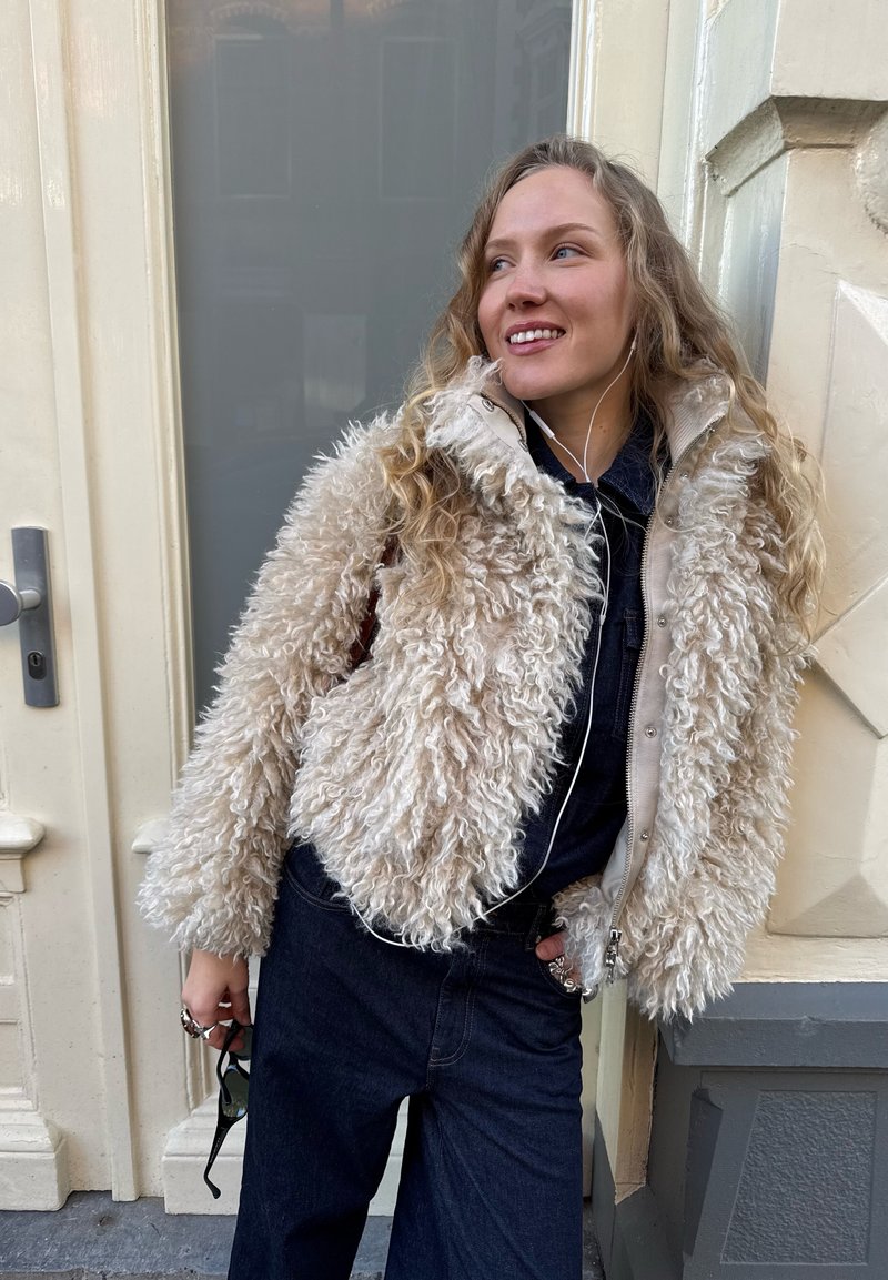 Jeune femme portant une veste beige duveteuse et un pantalon foncé, tenant des lunettes de soleil, debout près d'une porte avec des écouteurs.