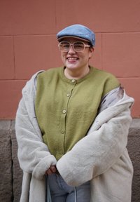 Χαμογελαστό άτομο που φοράει ανοιχτό γαλάζιο καπέλο, μεγάλα διαφανή γυαλιά, πουλόβερ χακί με κουμπιά και υπερμεγέθη μπεζ παλτό από συνθετική γούνα μπροστά από έναν τοίχο από τούβλα.