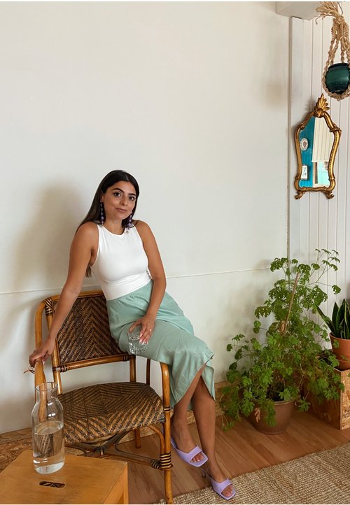 Egy fehér ujjatlan felsőt és menta zöld szoknyát viselő nő egy fonott székben ül, kezében egy pohárral, mellette egy üveg vízhűtő és cserepes növények vannak.