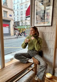 Gebreide groene turtleneck trui, gemêleerde grijze panterprintbroek, gezeten op een houten bank met een latte in een lichte kom, stedelijke caféomgeving.