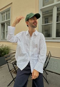 Camisa de algodón blanca con intrincados patrones de ojal, mangas largas y cierre de botones, combinada con pantalones de rayas azul marino y una gorra verde.
