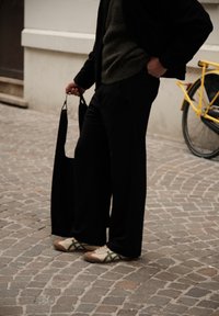 Zwarte wijde broek, olijfkleurige gebreide trui en casual beige schoenen met groene accenten. Een zwarte tas hangt aan de hand. Gele fiets op de achtergrond.