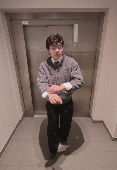 Joven con gafas que se ajusta los puños de la camisa frente a las puertas cerradas de un ascensor en un pasillo con azulejos.