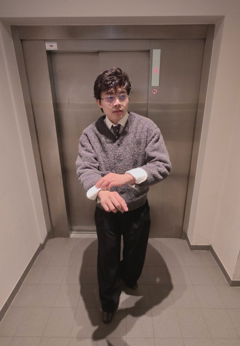 Jeune homme portant des lunettes ajustant les manches de sa chemise devant les portes d'ascenseur fermées dans un couloir carrelé.