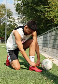 Mladý mužský futbalista kľačí na trávnatej ploche a viaže si jasne červené kopačky pri bielom a sivej futbalovej lopte vedľa kovového plotu.