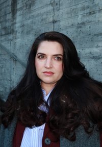 Mujer con cabello largo, oscuro y ondulado, vistiendo un abrigo gris sobre una camisa con rayas azules, posando frente a una pared de concreto texturizada.