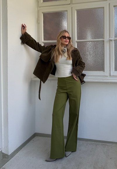 Mujer con gafas de sol, camiseta blanca, pantalones verde oliva y chaqueta marrón oscuro apoyada contra una pared blanca cerca de ventanas esmeriladas.