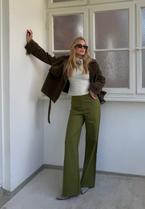 Femme portant des lunettes de soleil, un haut blanc, un pantalon vert olive et une veste marron foncé, appuyée contre un mur blanc près de fenêtres givrés.