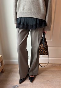 Světle šedé kalhoty s širokými nohavicemi, černé boty s špičatou špičkou, navrstvené s černým topem s krajkovým lemem, držící kabelku s leopardím vzorem.