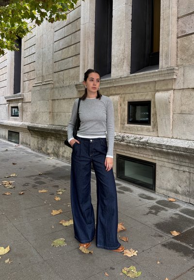 Top de manga larga gris superpuesto sobre blanco, combinado con pantalones de mezclilla de pierna ancha en azul oscuro. De pie en una acera con hojas de otoño esparcidas.