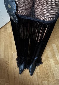 Ben klædt i sorte fiskenet-strømper, frynseskørt og spidse sorte læderstøvler på trægulv.